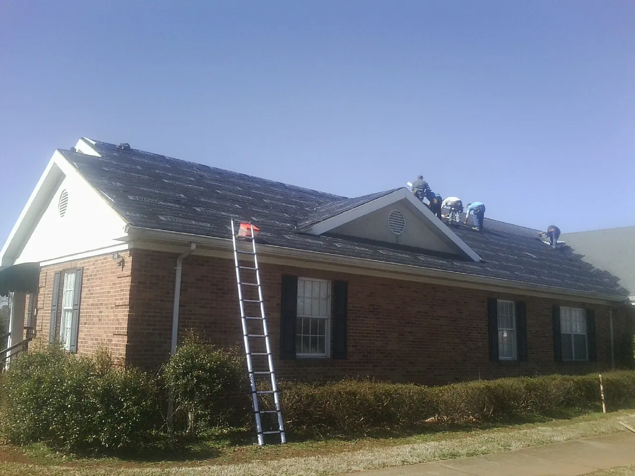 Crew tearing shingles off a brick ranch roof with a ladder set — Metal Roof Repair work by our crew in Martin City