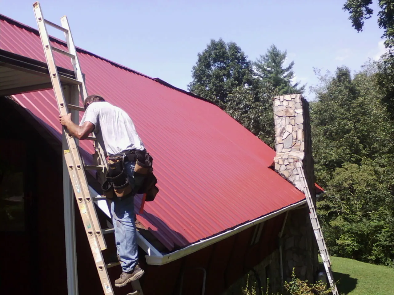 Roofer on a ladder beside a red metal roof and stone chimney — Soffit & Fascia Repair by Roofing Brothers in Martin City
